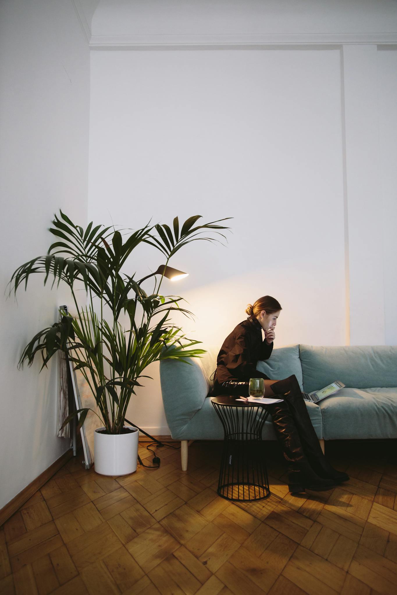 A woman sits on a sofa with a laptop, surrounded by indoor plants in a modern living room setting.
