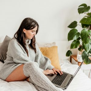 Young woman in cozy sweater, using laptop on bed with plant nearby.